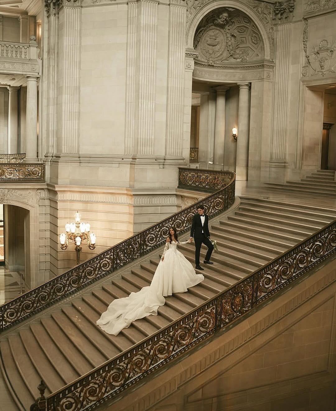 Bride and groom walking down a grand stone staircase in a classical hall.