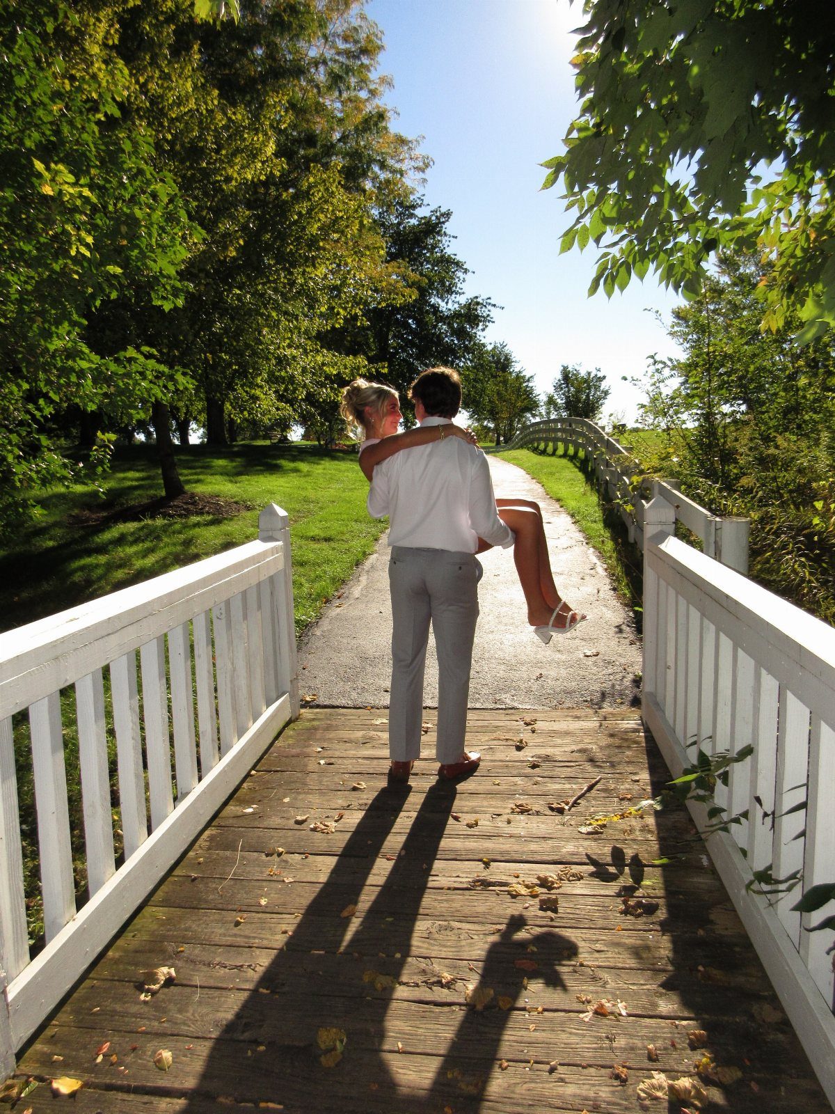 Man lifting woman in white dress on a wooden bridge surrounded by trees