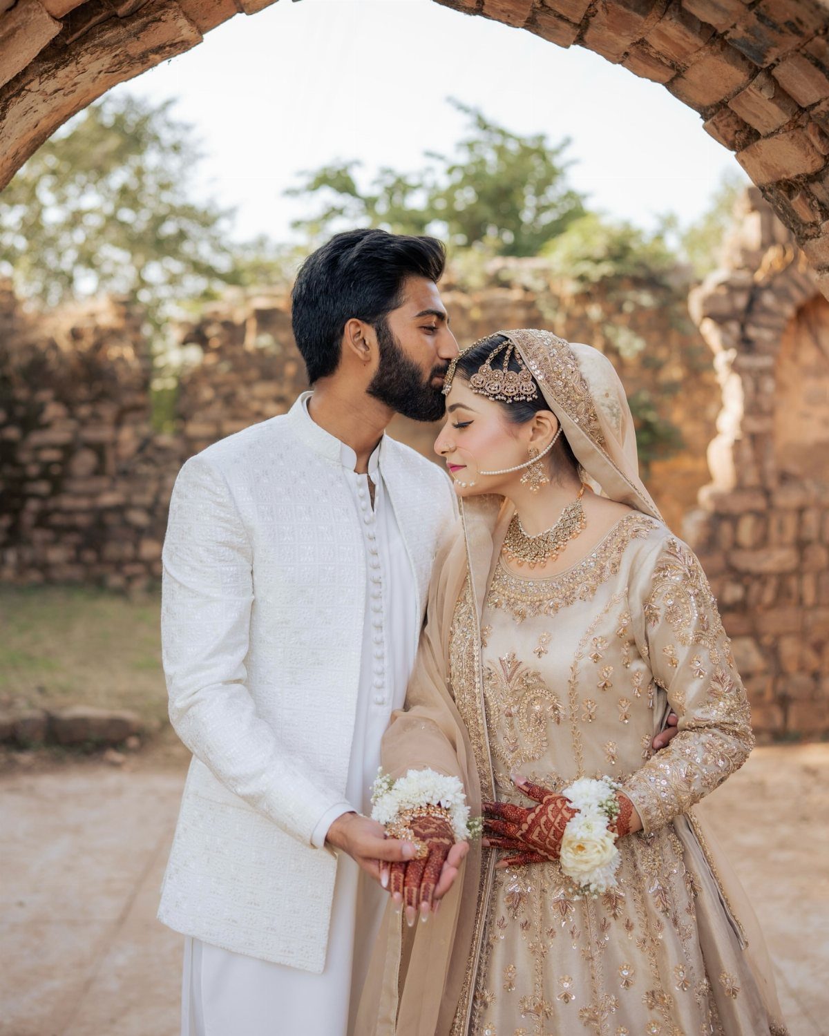 Groom in white sherwani kissing bride's forehead under a rustic stone archway