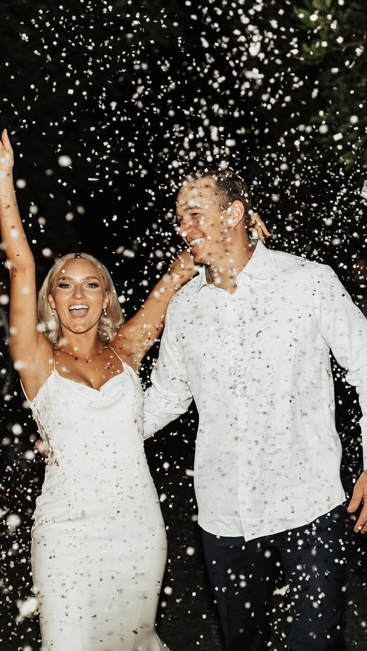 Smiling wedding couple celebrating under a shower of falling white confetti at night.