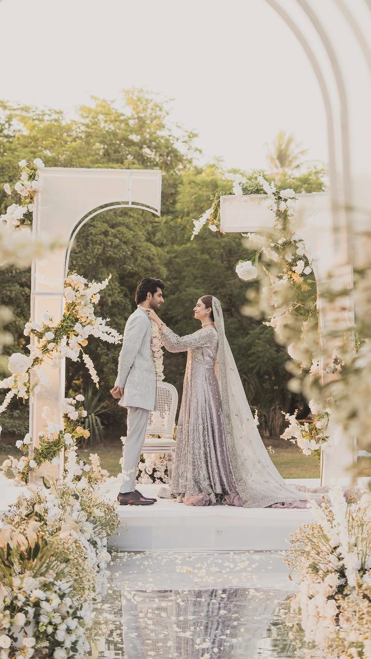 Bride and groom facing each other under a white floral arch in a garden