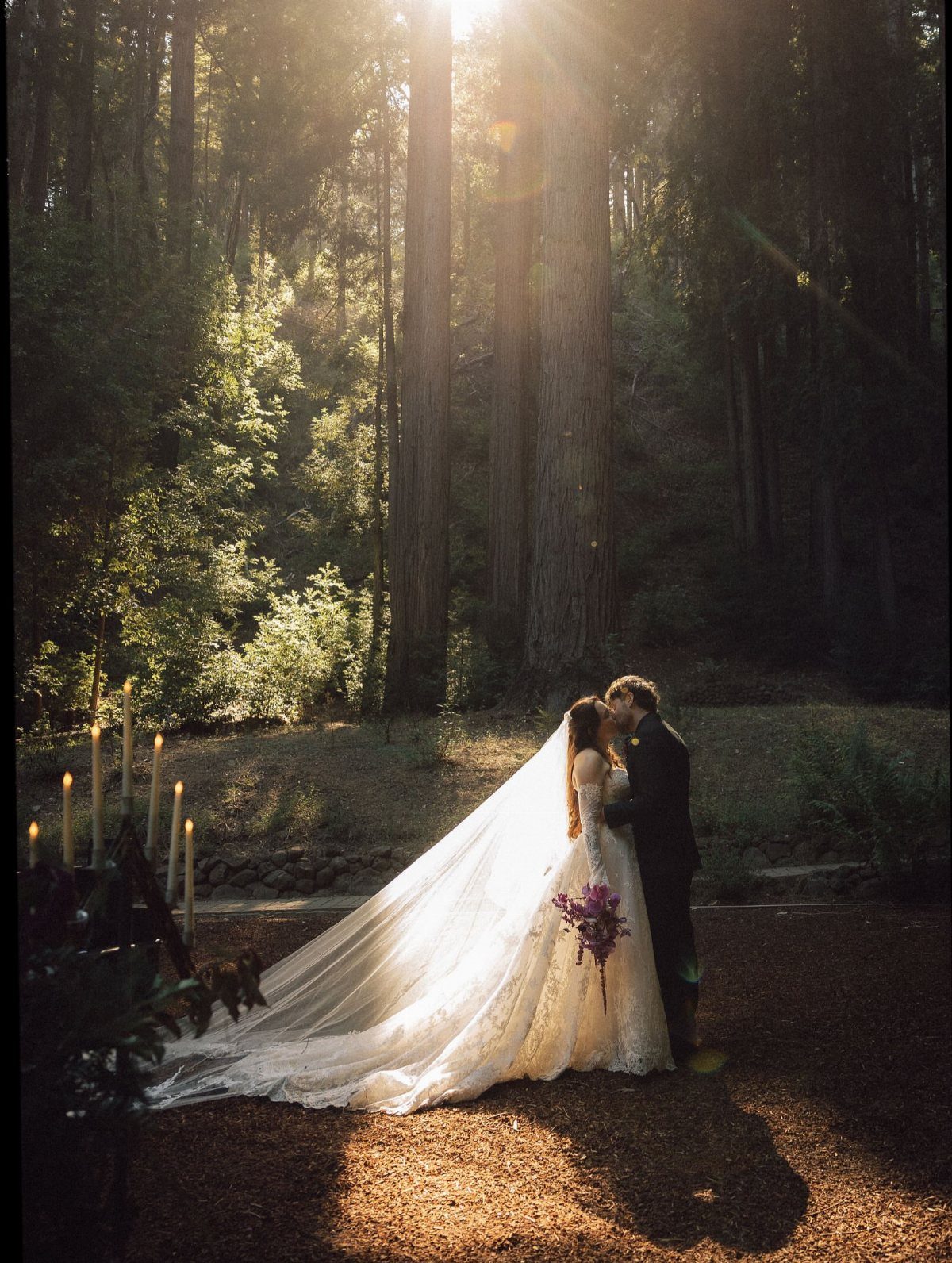 Bride and groom kissing in a sunlit redwood forest with lit candles nearby.