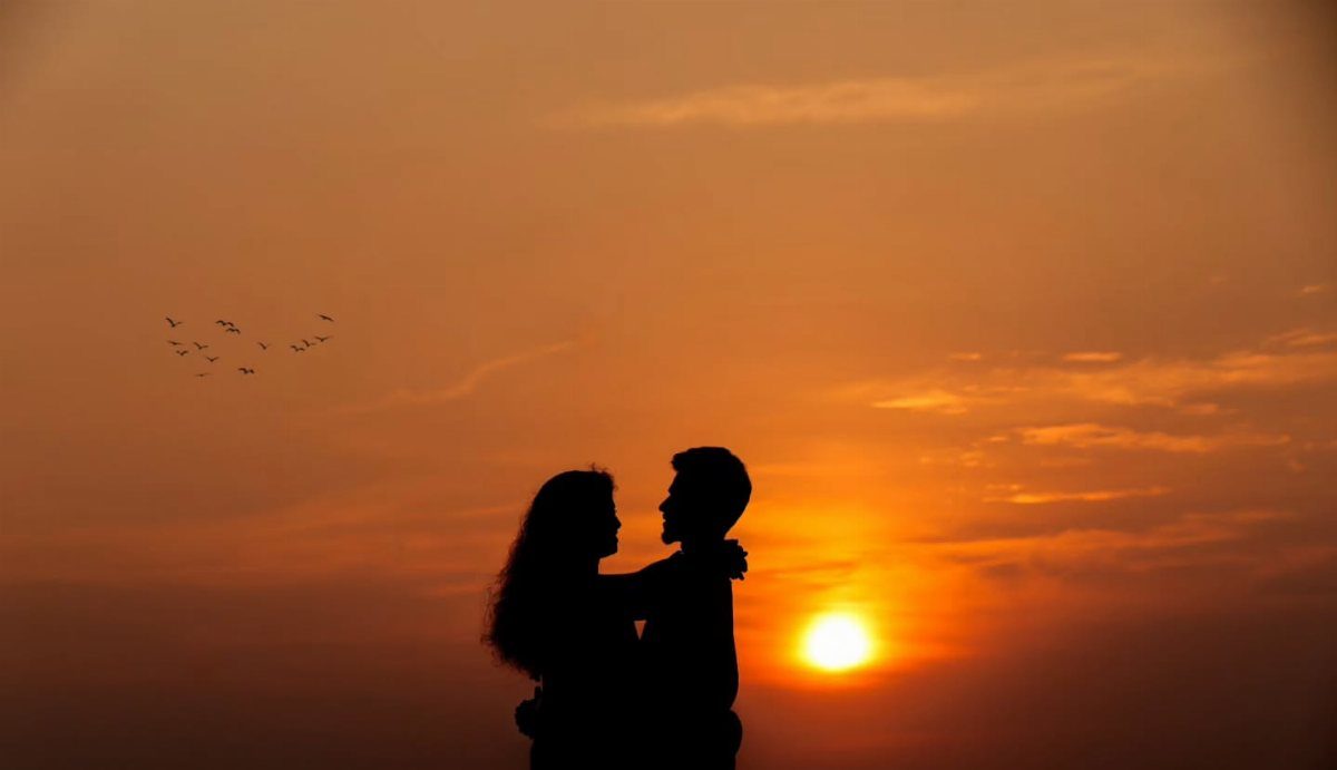 Couple in silhouette embracing against a bright orange sunset sky with flying birds.