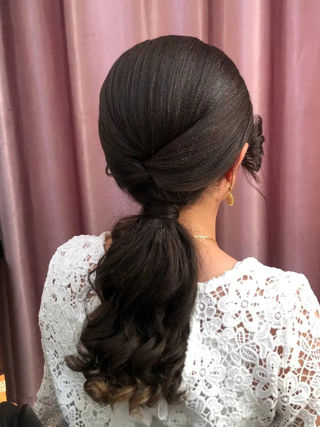 Dark brown hair styled in a curly low ponytail with a white lace dress.