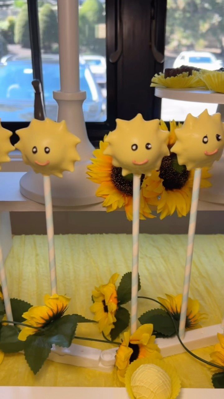 Sun-shaped yellow cake pops with smiling faces displayed on a white stand with sunflowers.