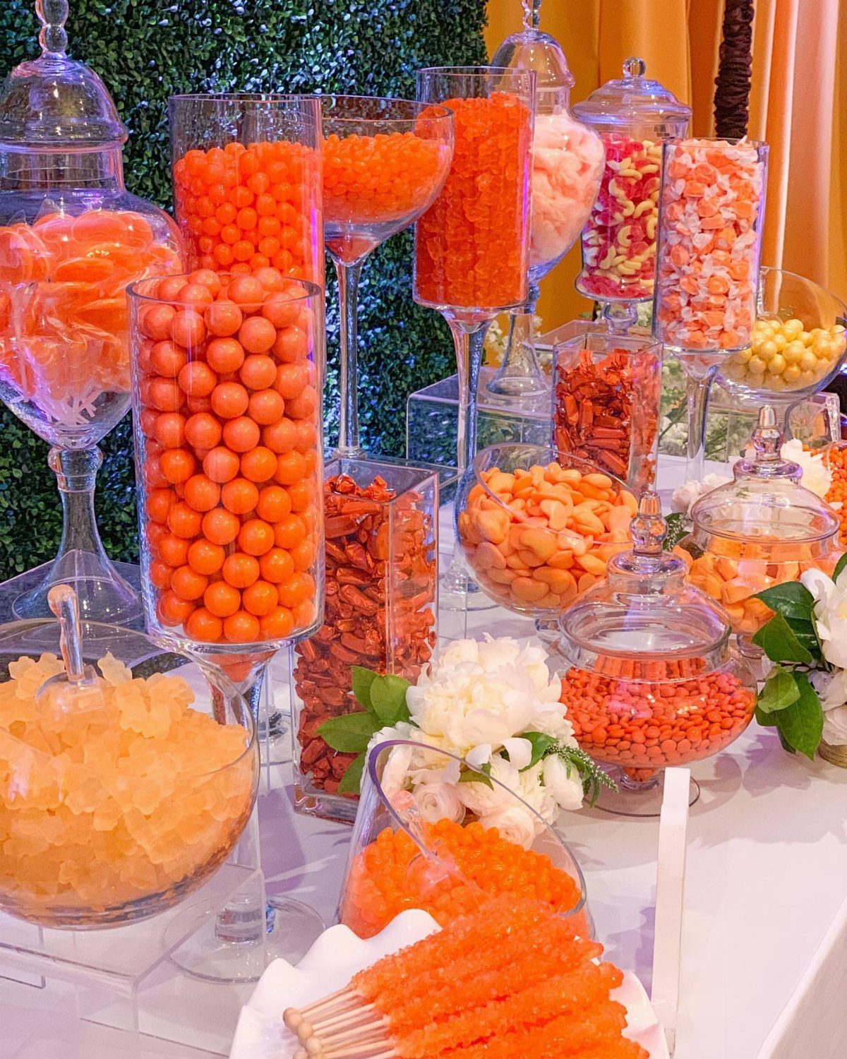 Assorted orange and pink candies in glass jars on a white wedding dessert table.