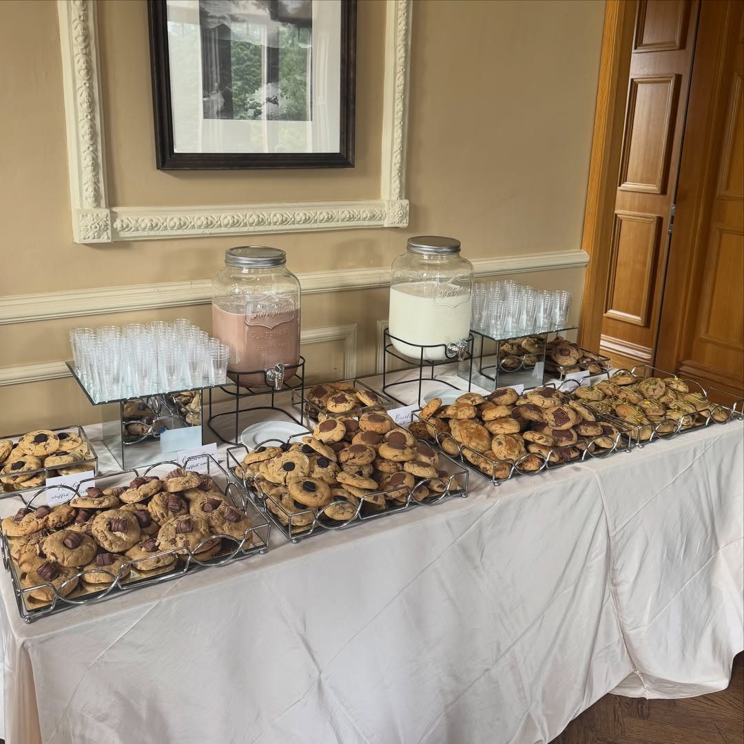 Cookie bar with trays of chocolate chip cookies and milk dispensers on white tablecloth.