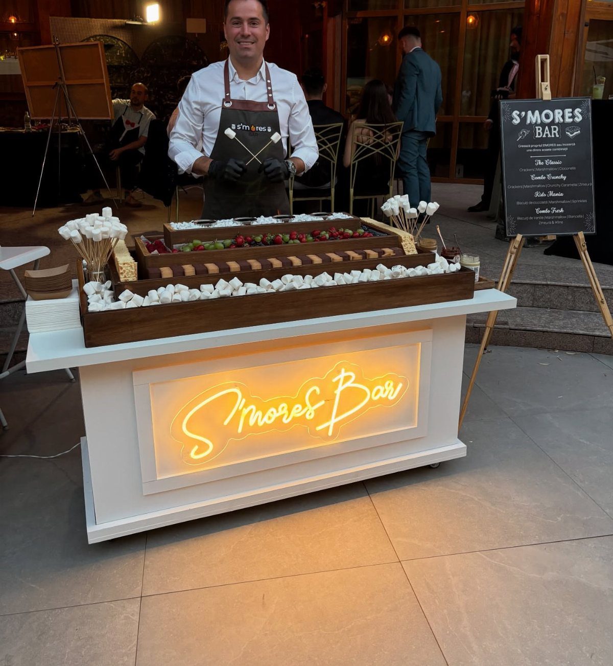 S'mores bar station with marshmallows and chocolate, attended by a man in an apron.