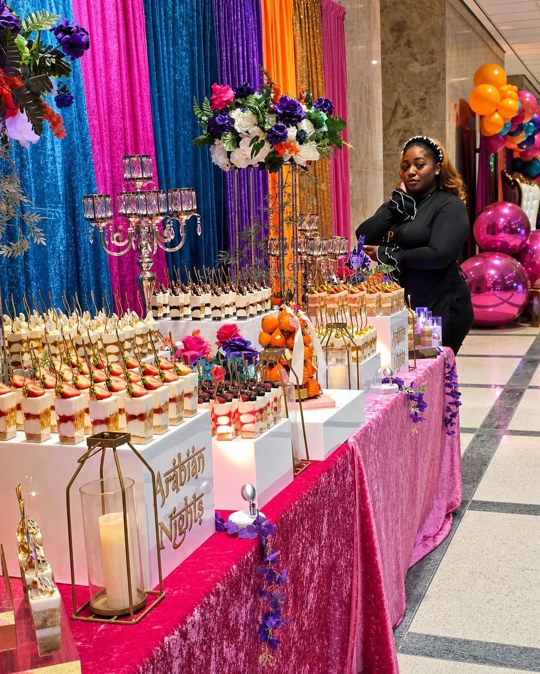Dessert table with pink velvet cloth and colorful curtains featuring various miniature cakes.