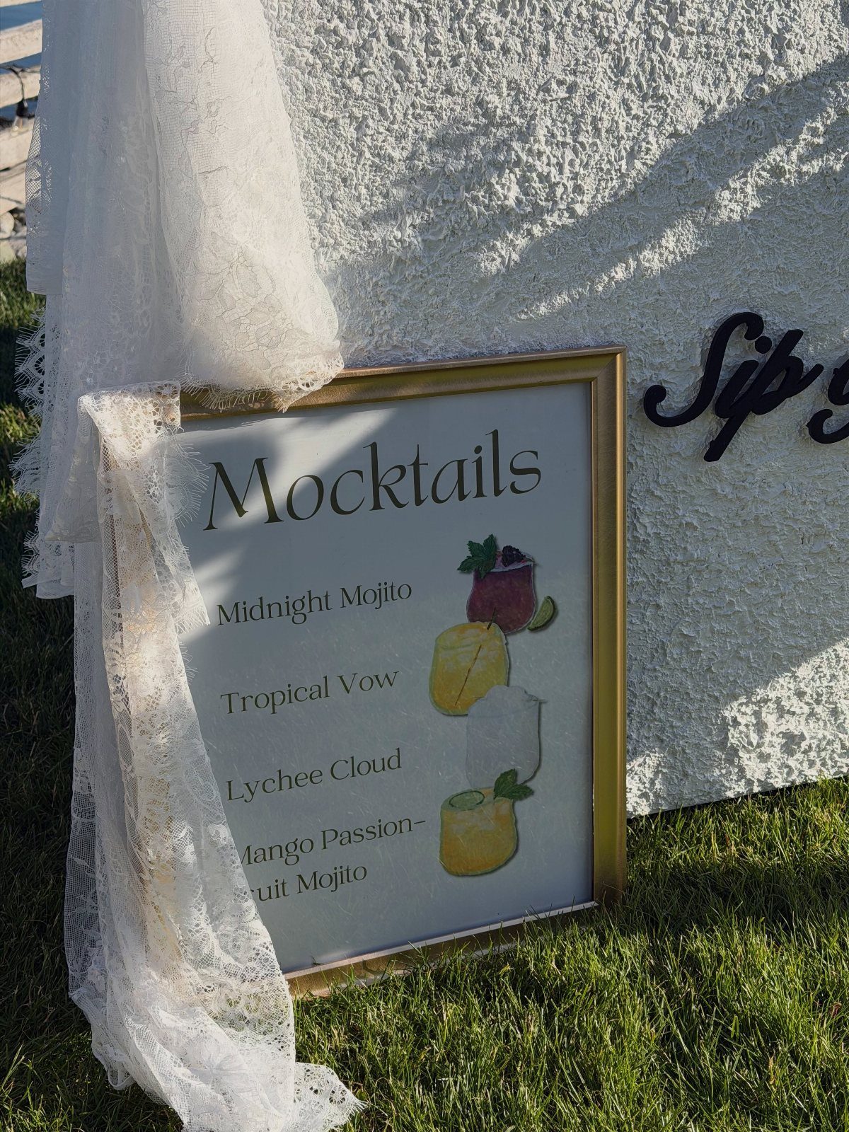 Mocktail menu sign in a gold frame draped with white lace on grass.