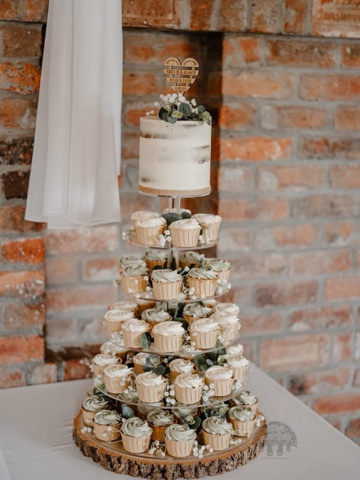 Semi-naked wedding cake atop a tiered stand filled with cupcakes on a wooden base.