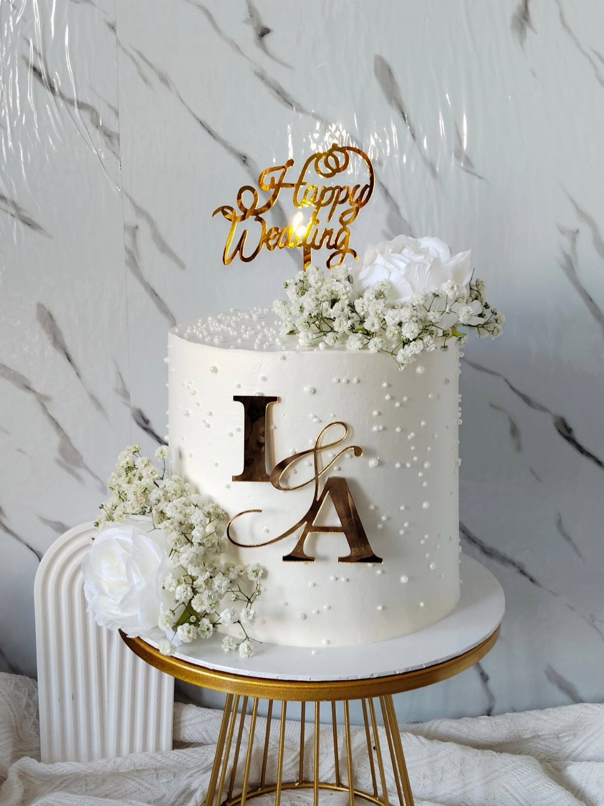 Single-tier white wedding cake with gold toppers and white floral decorations on gold stand