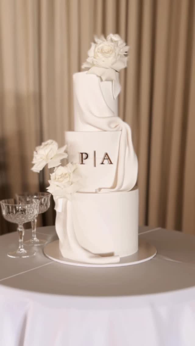 Three-tier white wedding cake with draped fondant, white flowers, and gold initials.