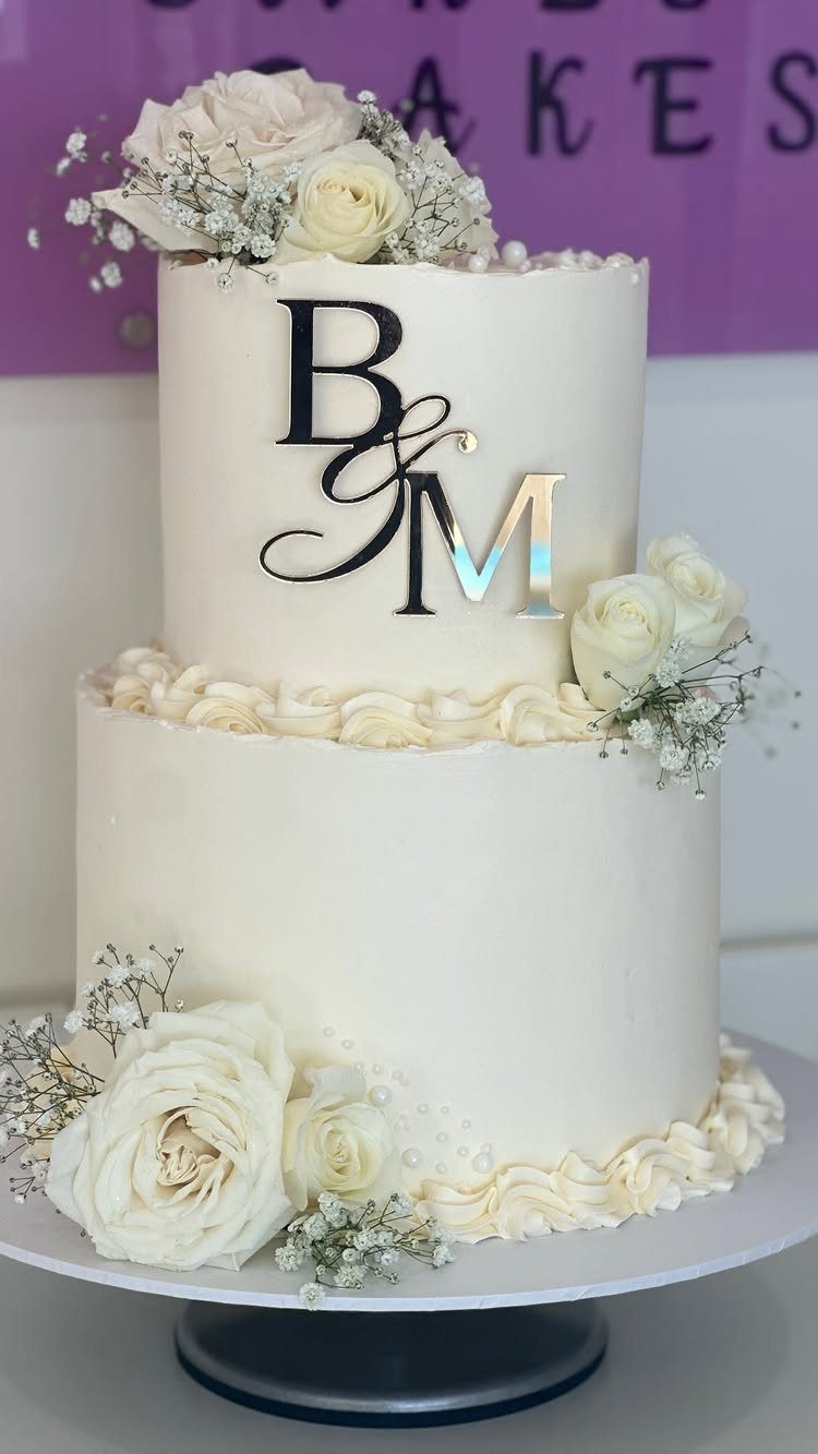 Two-tier white wedding cake decorated with white roses and a silver monogram topper.
