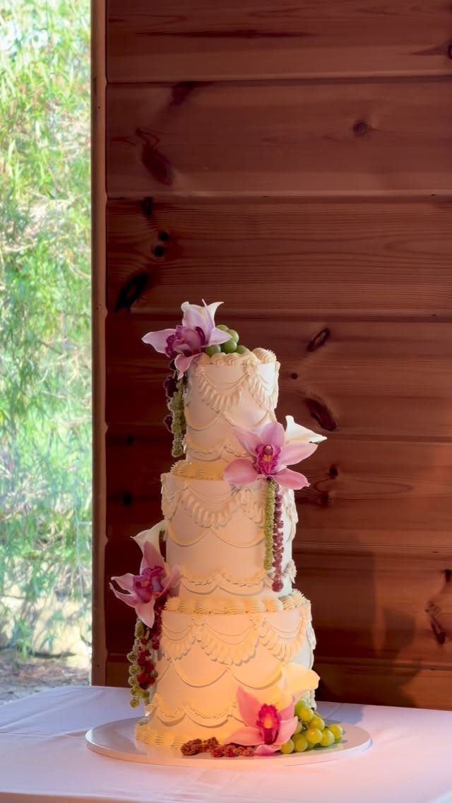 Three-tier white wedding cake with scalloped icing, purple orchids, and green grapes.
