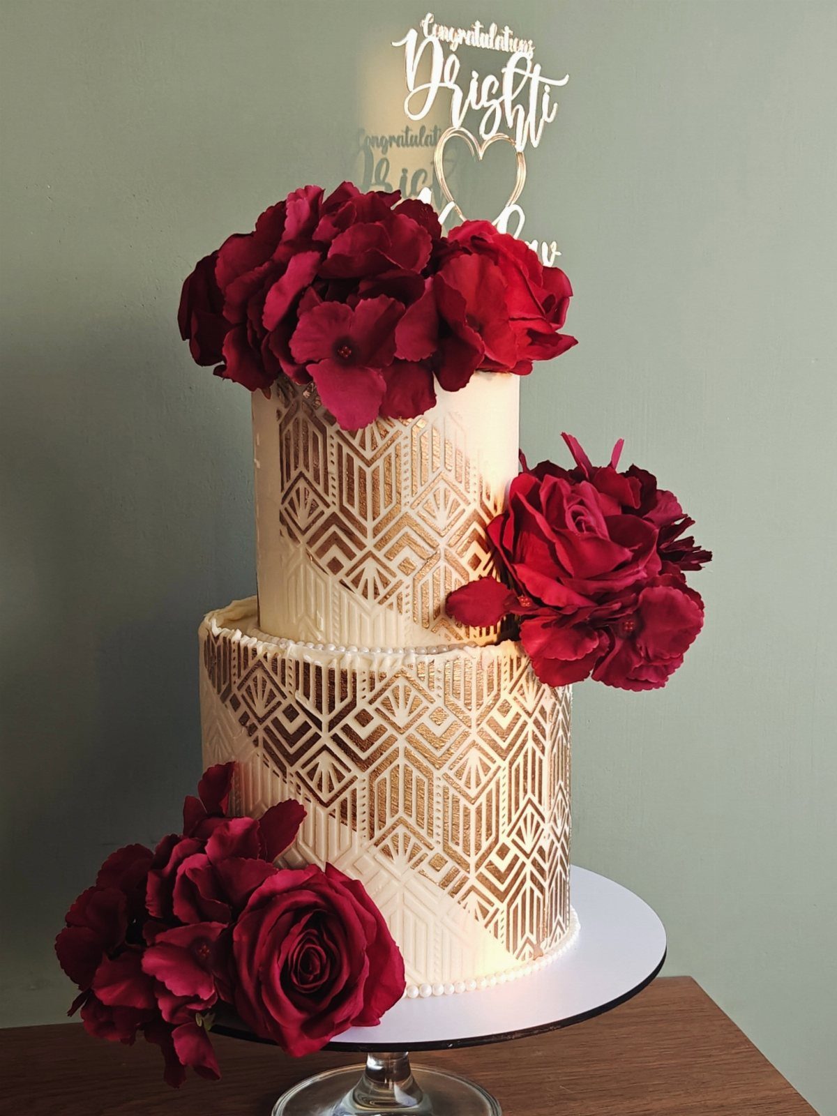 Two-tier white cake with gold geometric patterns and red roses on a stand.