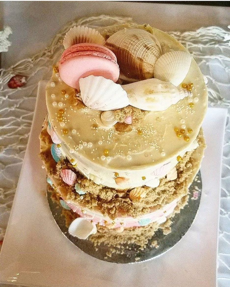 Beach-themed cake decorated with seashells, sand crumbs, and a pink macaron.