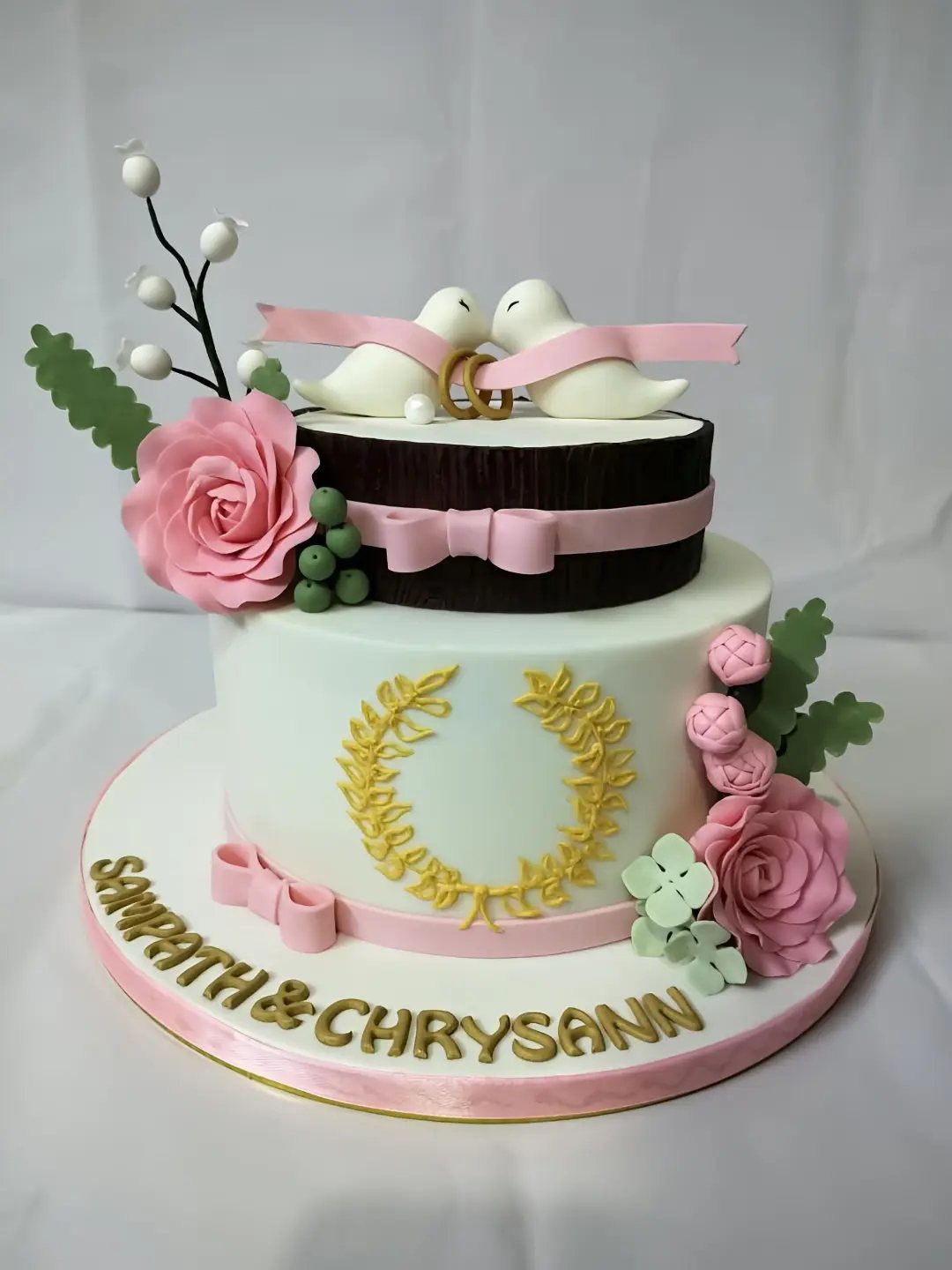 Two-tier engagement cake with white bird figurines, pink roses, and gold lettering.