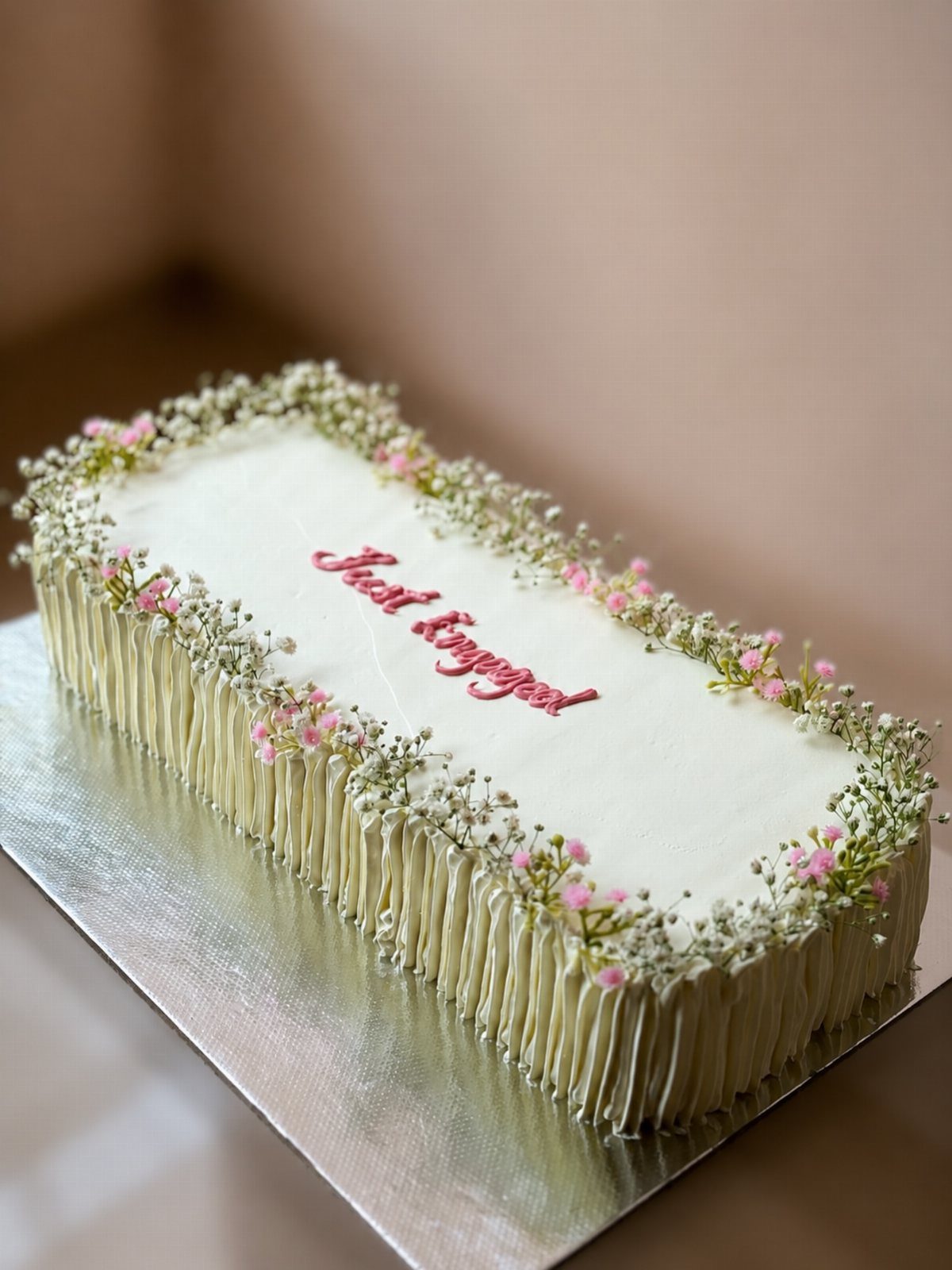 Rectangular white engagement cake decorated with delicate pink and white flowers and red script.