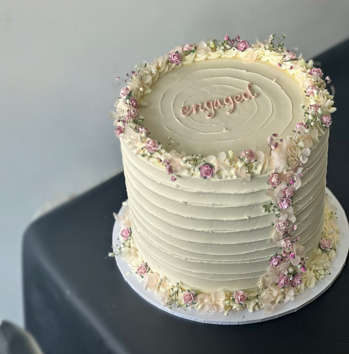 White ridged cake decorated with small pink flowers and 'engaged' written on top.