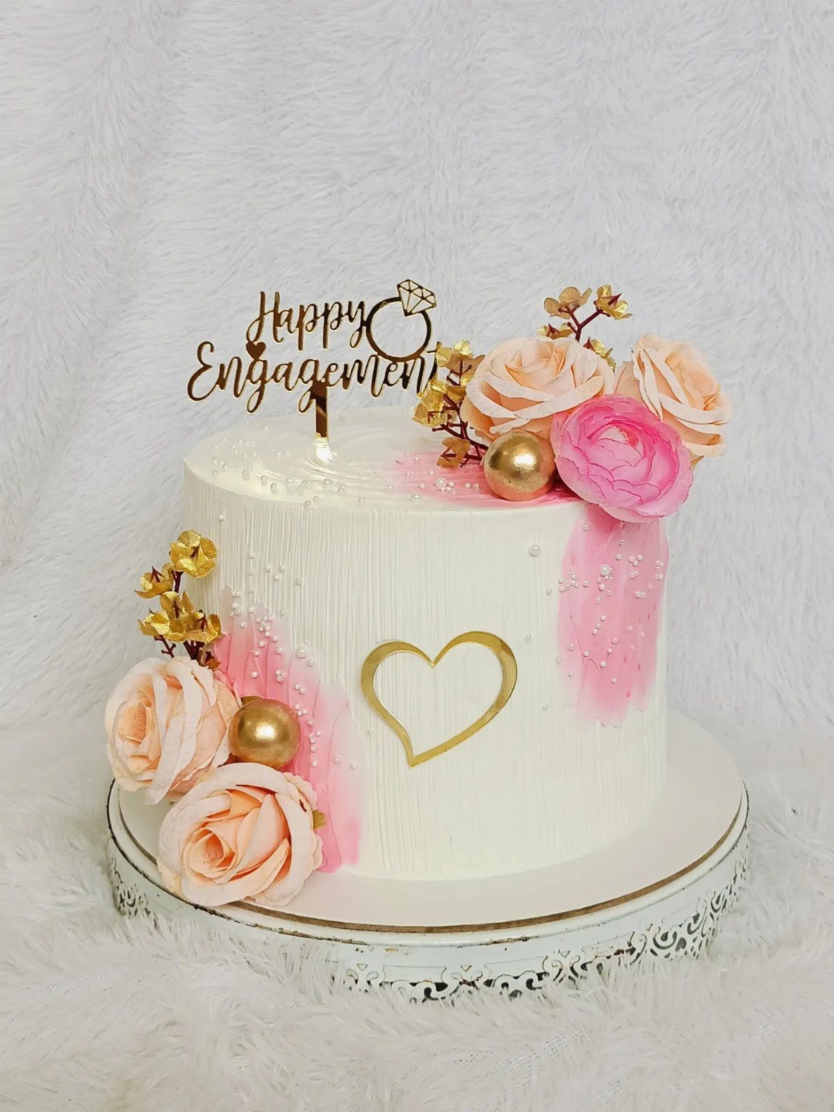 White engagement cake with pink accents, gold heart, and peach roses on stand.