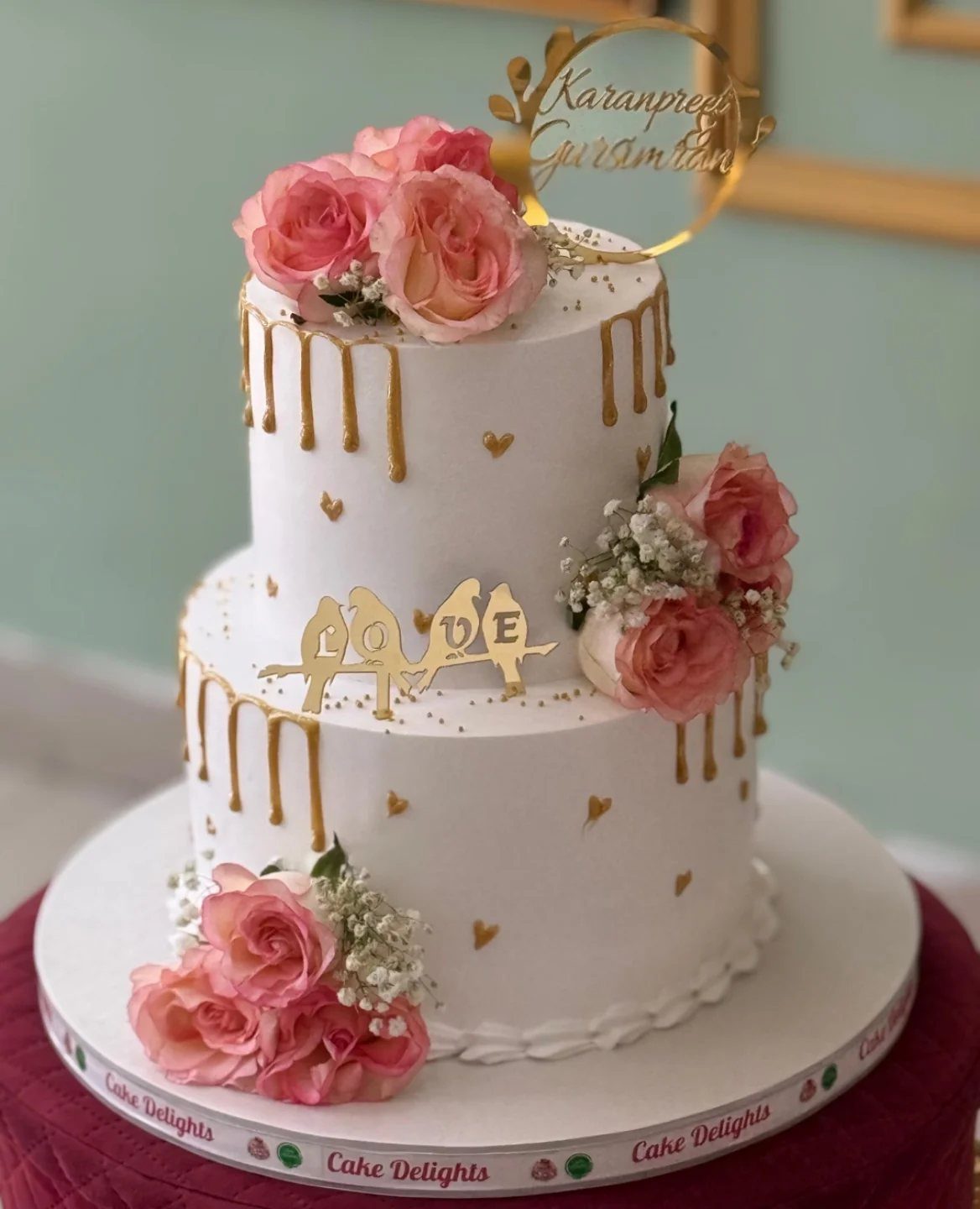 Two-tier white cake with golden drips, pink roses, and gold love topper.