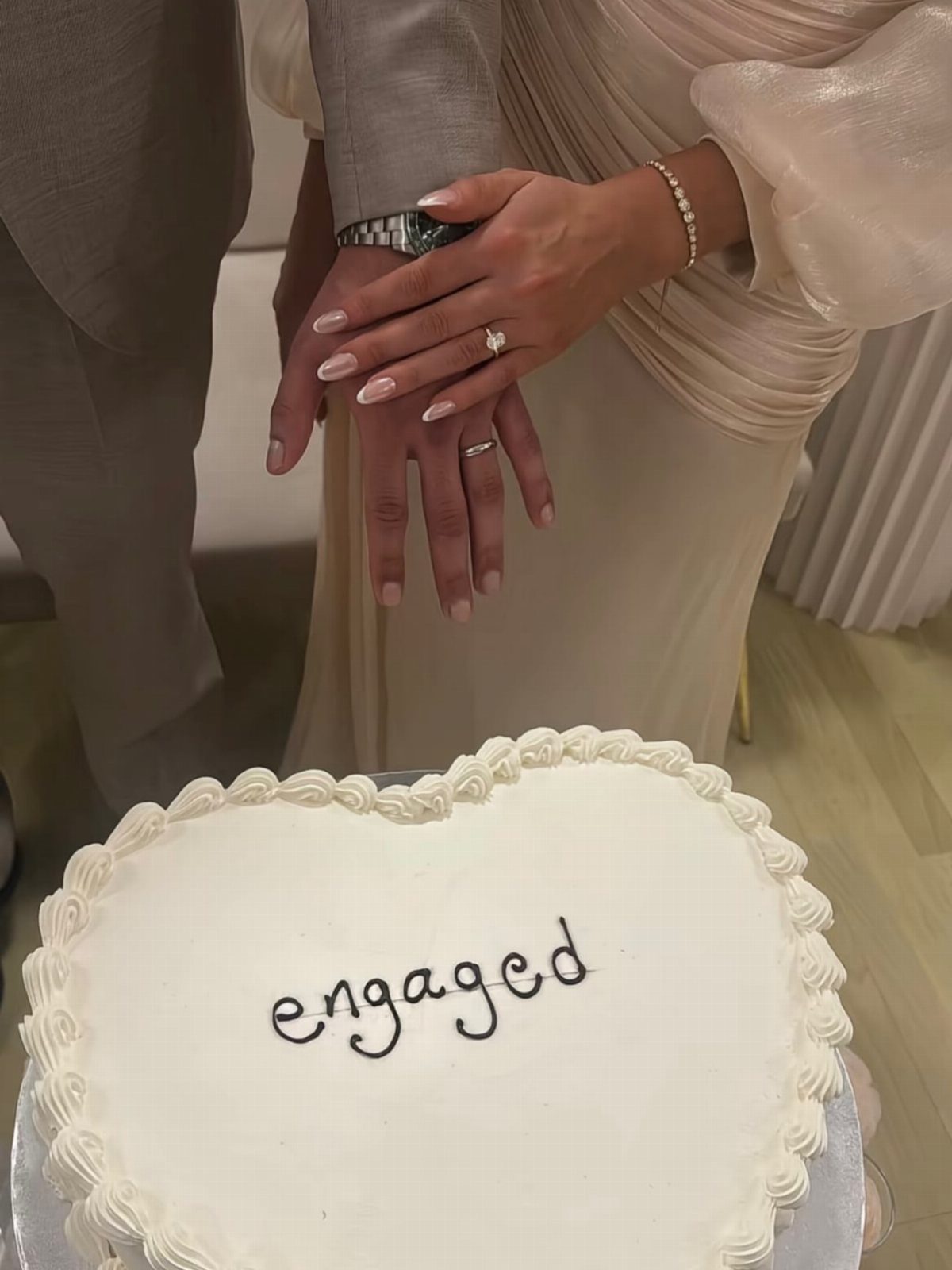 Heart-shaped white cake with engaged written on top and couple's hands above.