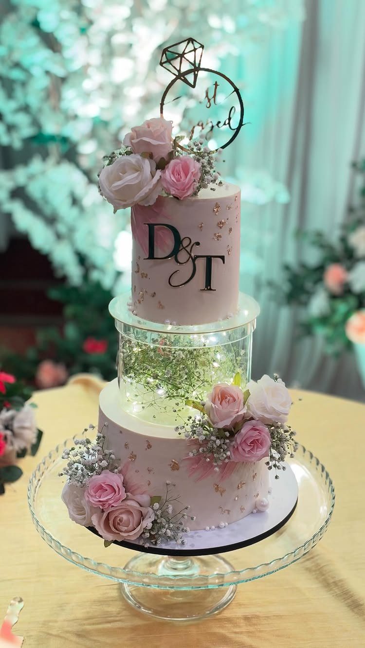 Three-tier pink engagement cake with roses, fairy lights, and a diamond ring topper.