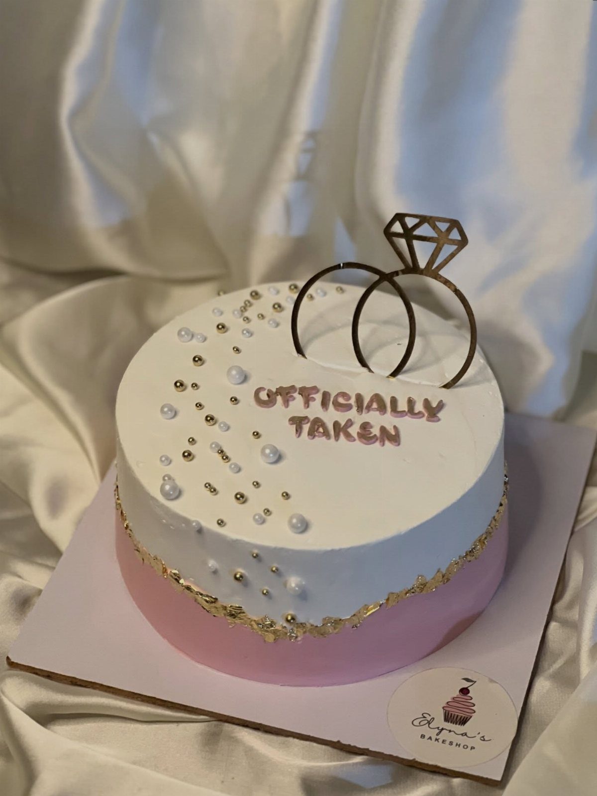 White and pink engagement cake with gold ring topper and 'Officially Taken' text.