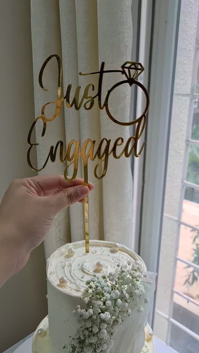 White frosted cake with baby's breath and a gold Just Engaged topper.