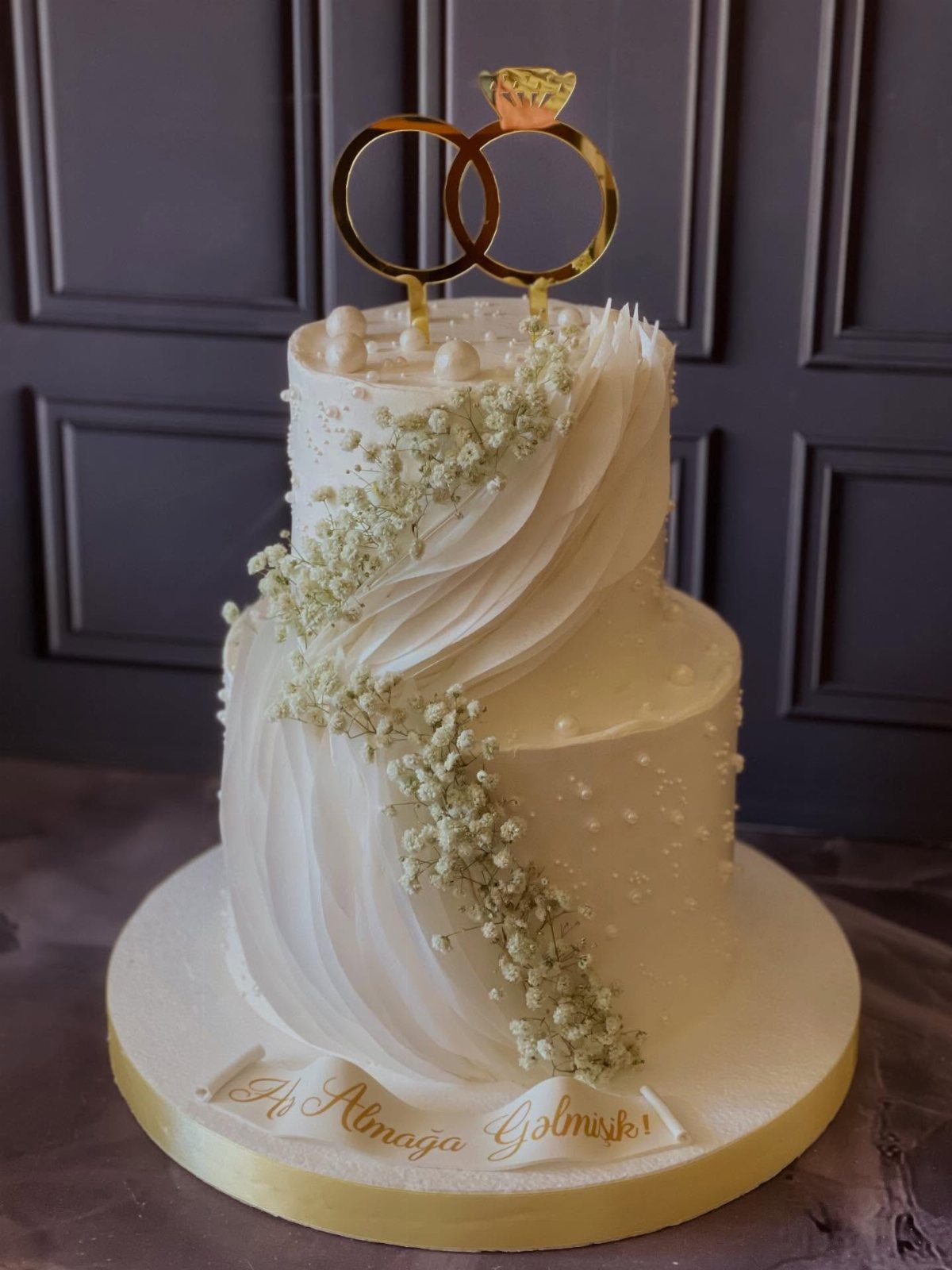 Two-tier white cake with cascading baby's breath flowers and gold ring topper.