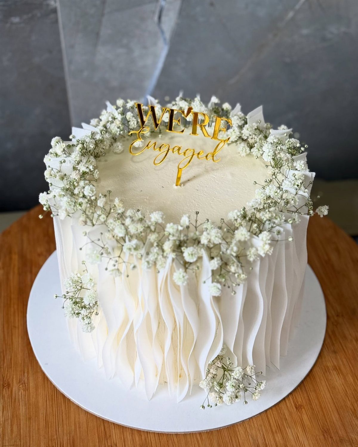 White ruffled cake topped with baby's breath flowers and gold engagement topper.