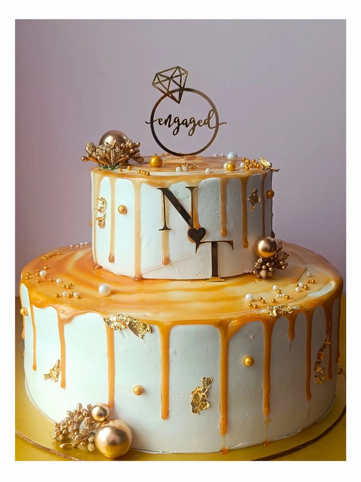 Two-tier white cake with golden caramel drip, gold pearls, and engaged ring topper.