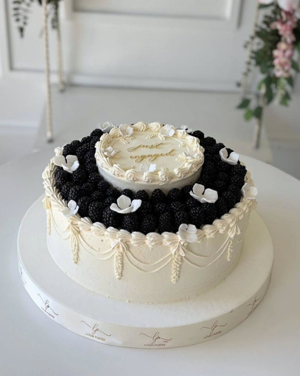 Two-tier white cake decorated with blackberries, white flowers, and gold script writing.