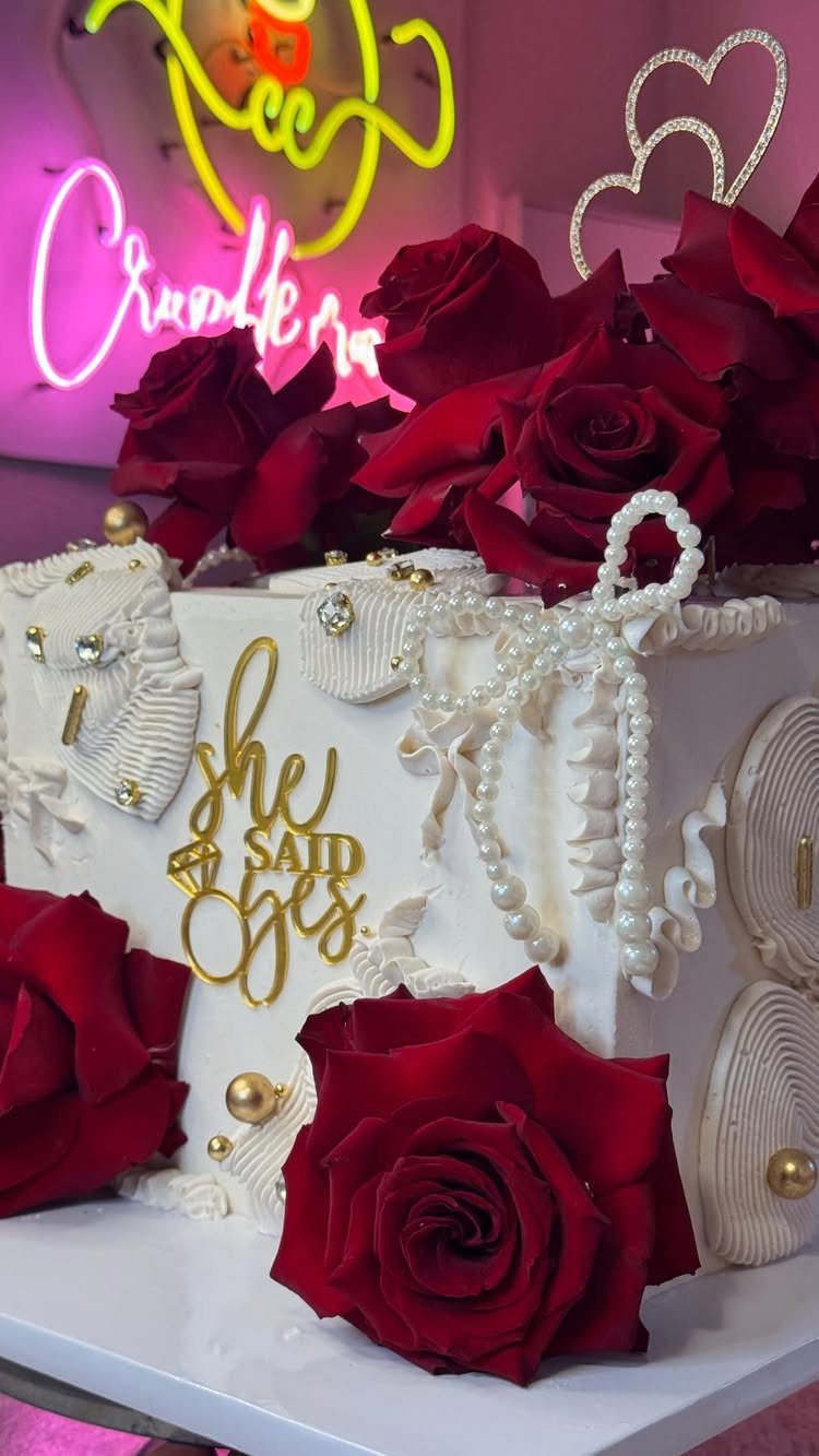 White engagement cake decorated with red roses and a gold she said yes topper