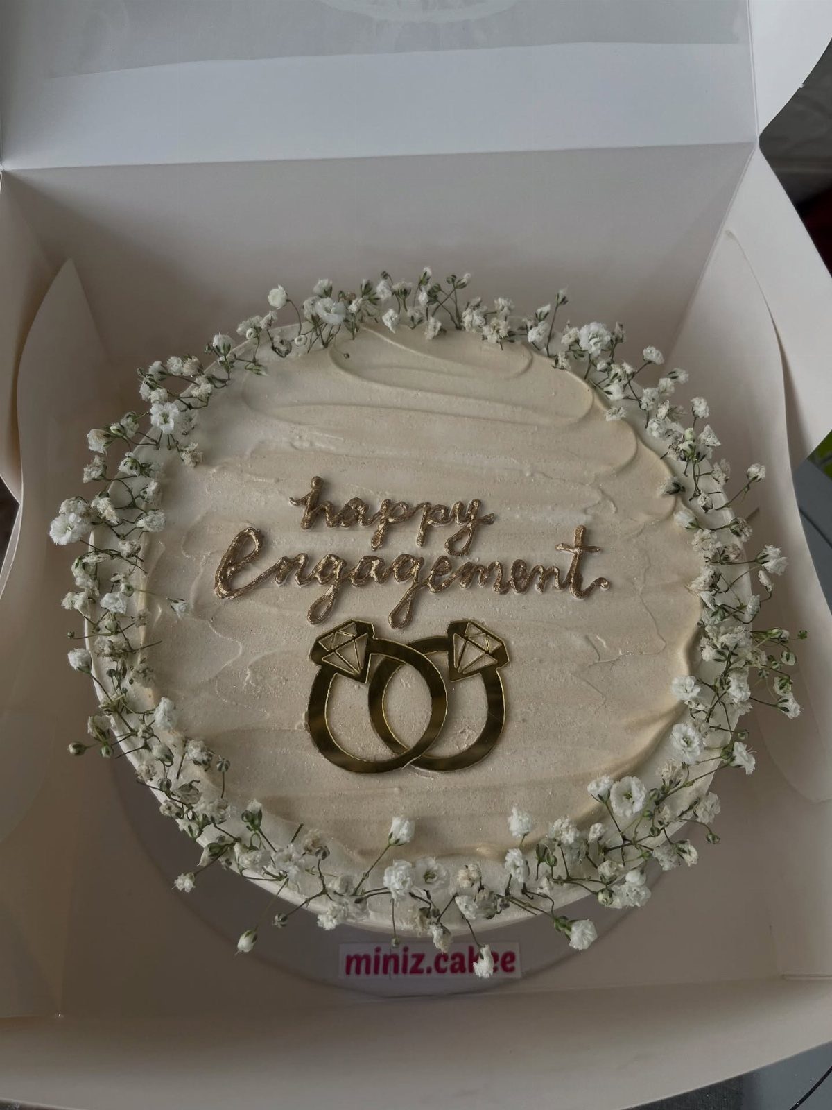 White frosted engagement cake with baby's breath flowers and gold ring toppers.