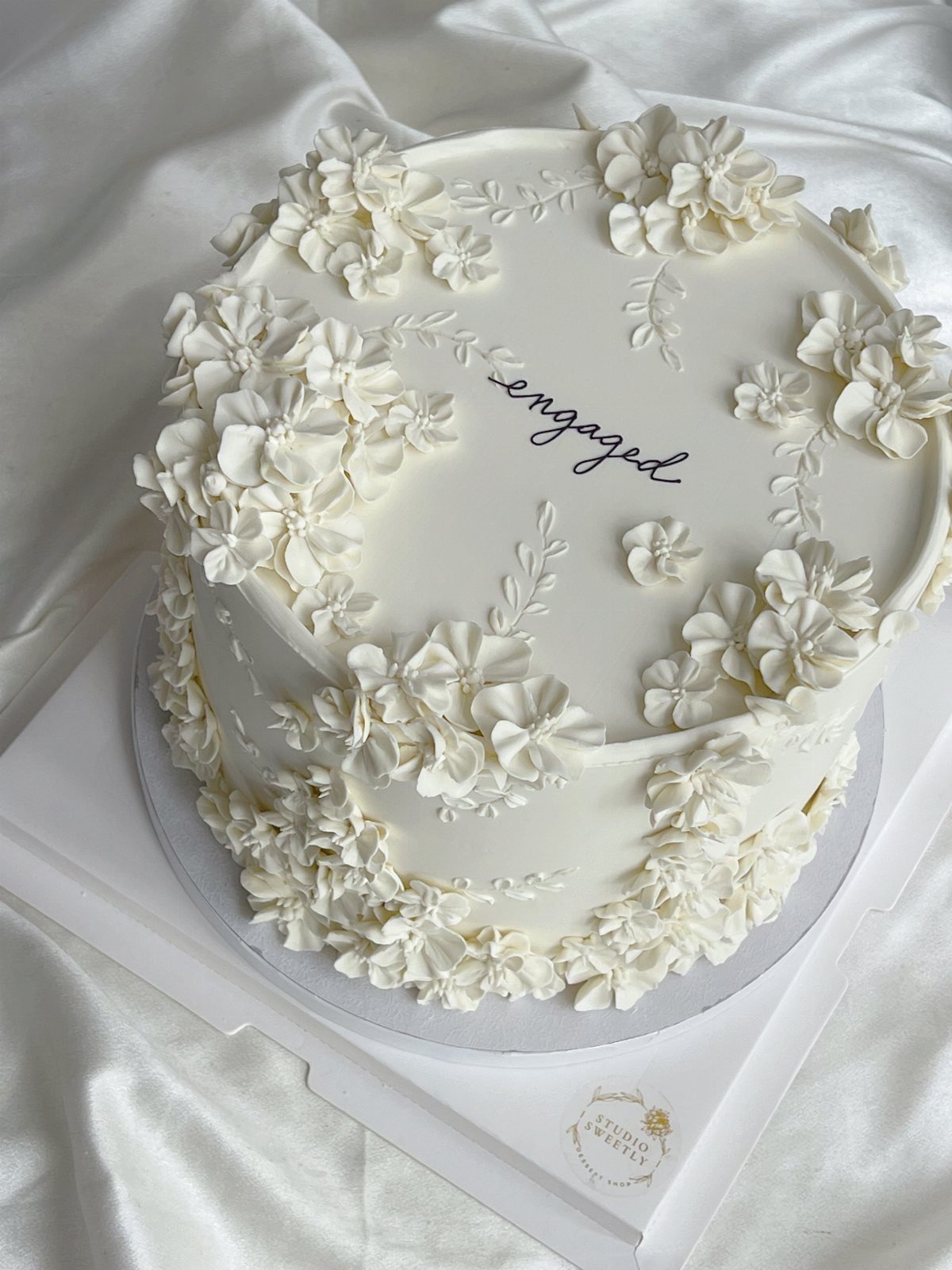 White engagement cake decorated with buttercream flowers and the word engaged in cursive.