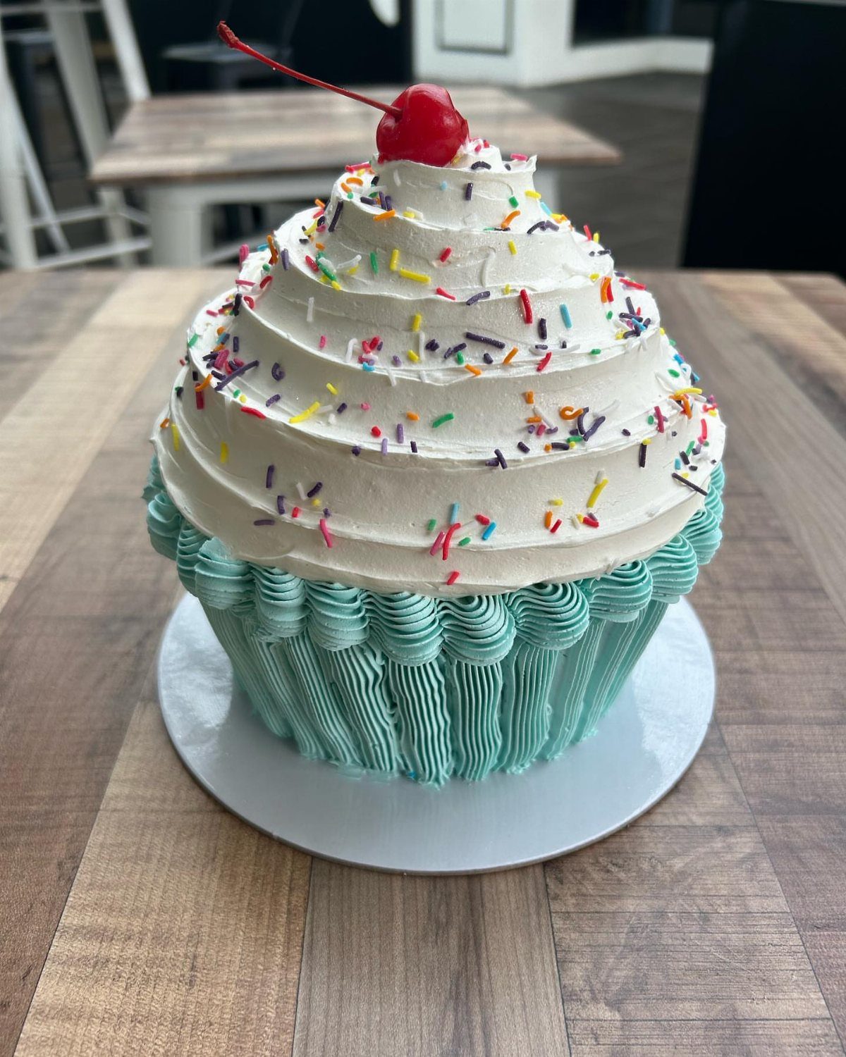 Giant cupcake cake with teal frosting base, white swirls, sprinkles, and a cherry.