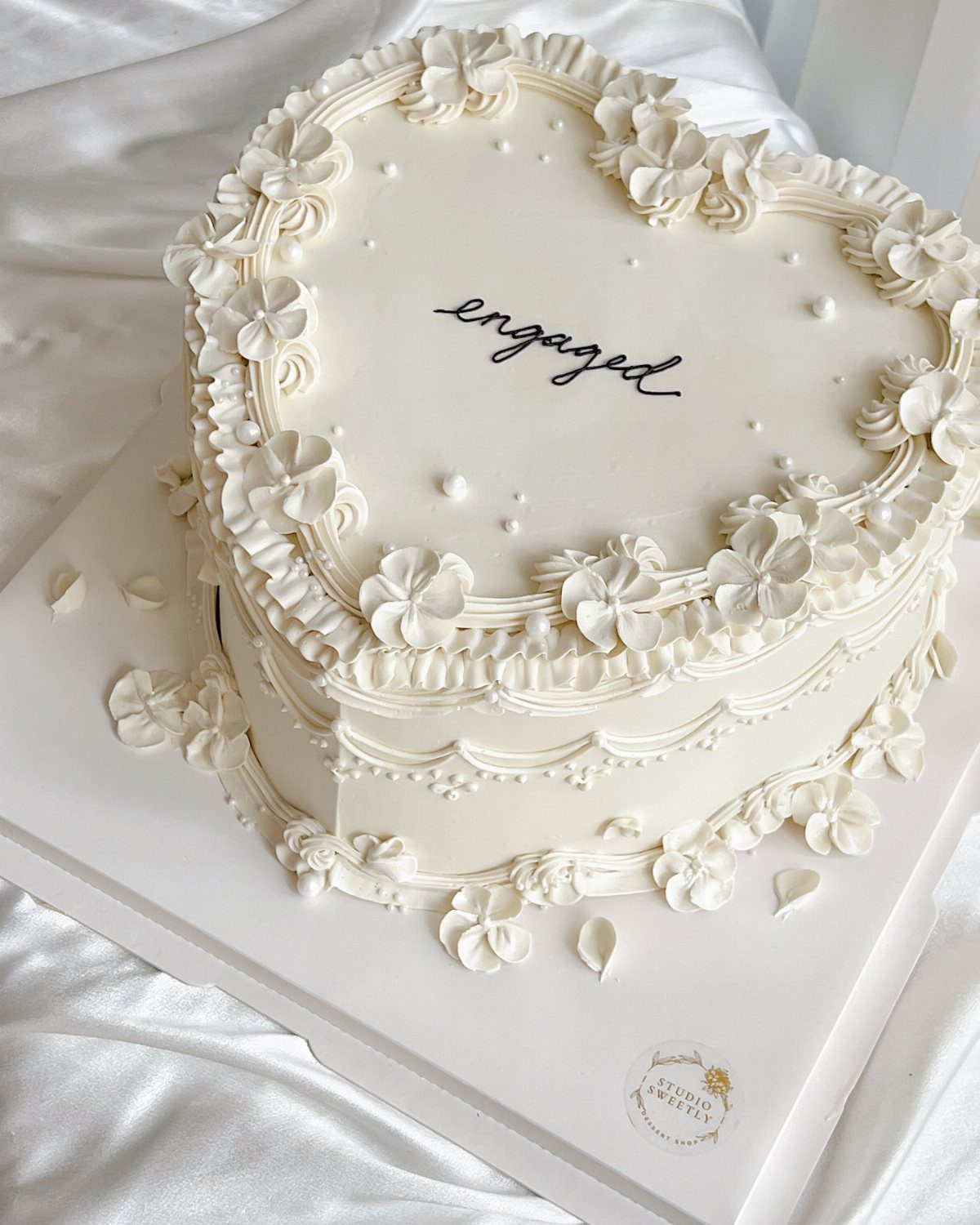 Heart-shaped white engagement cake decorated with piped cream flowers and black cursive text.