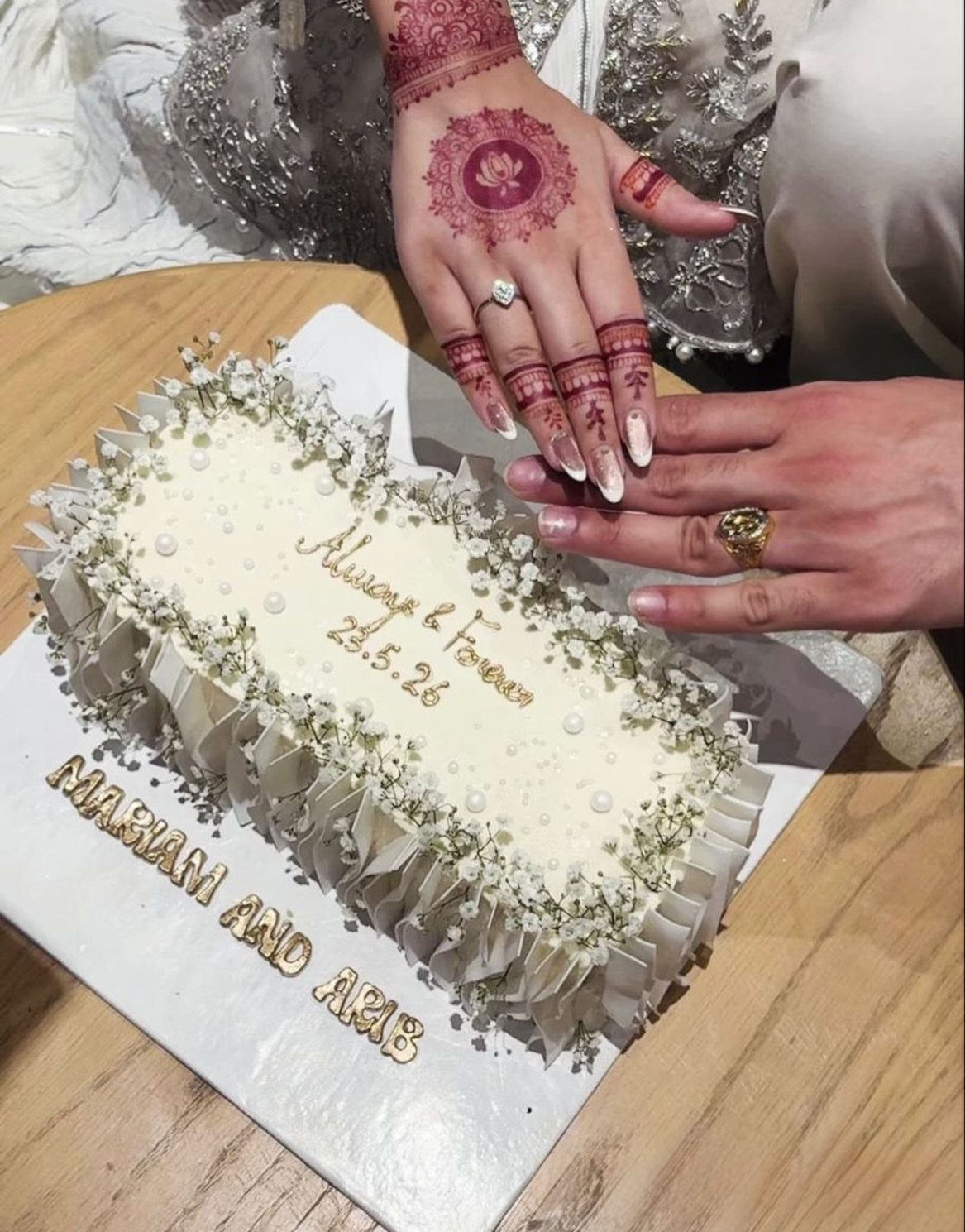 White rectangular cake with envelopes and flowers, with hands wearing rings above it.