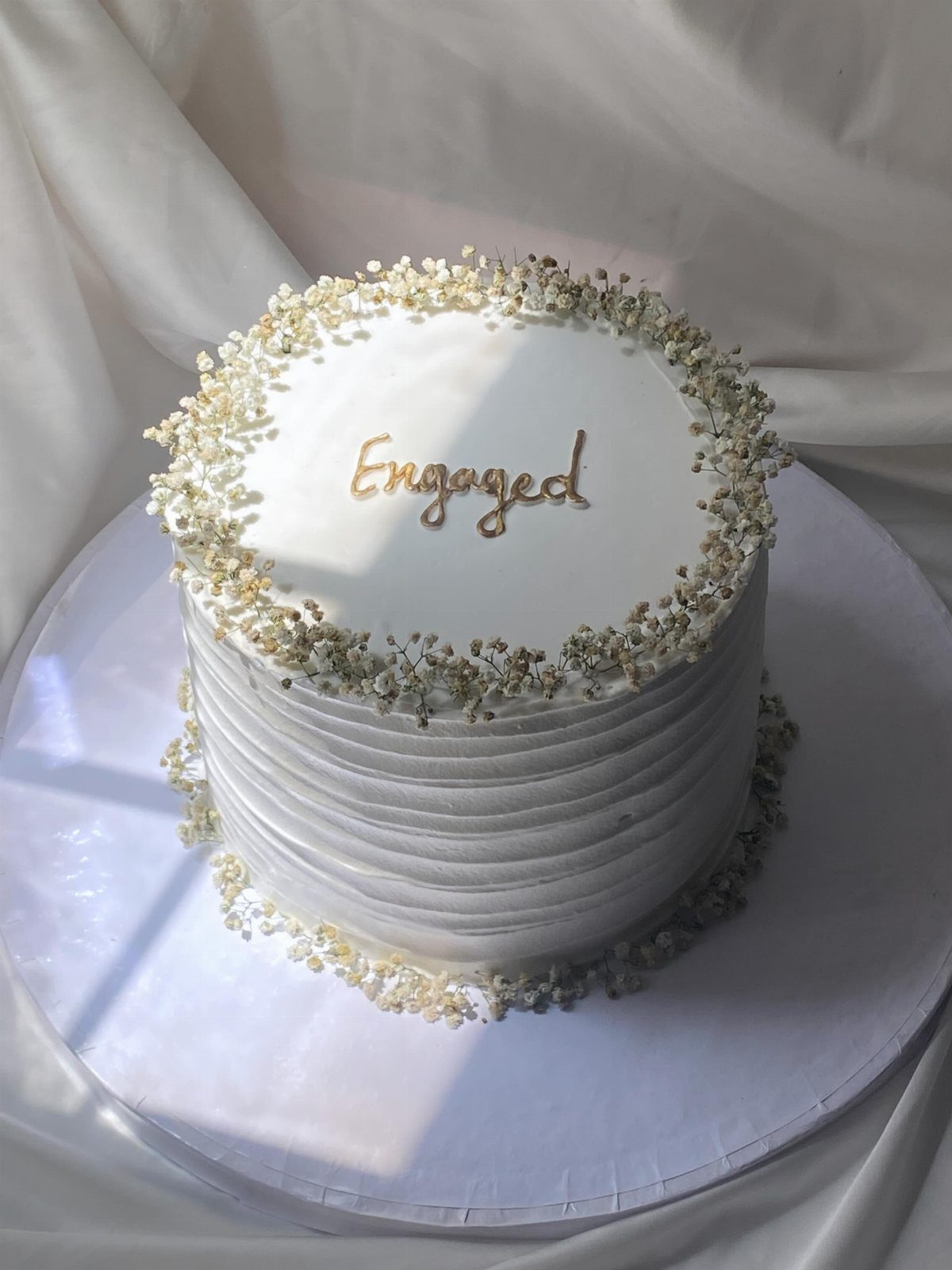 White frosted engagement cake decorated with baby's breath and gold Engaged topper.