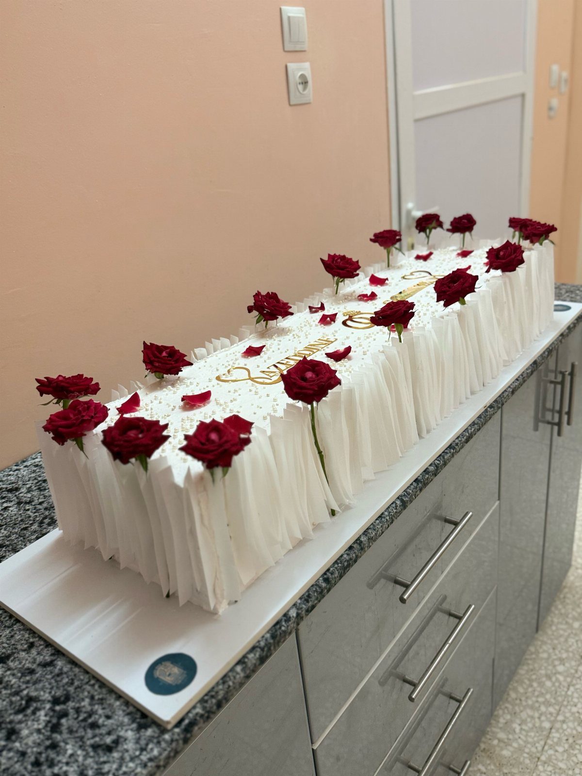 Long white rectangular cake decorated with red roses and gold cursive lettering.