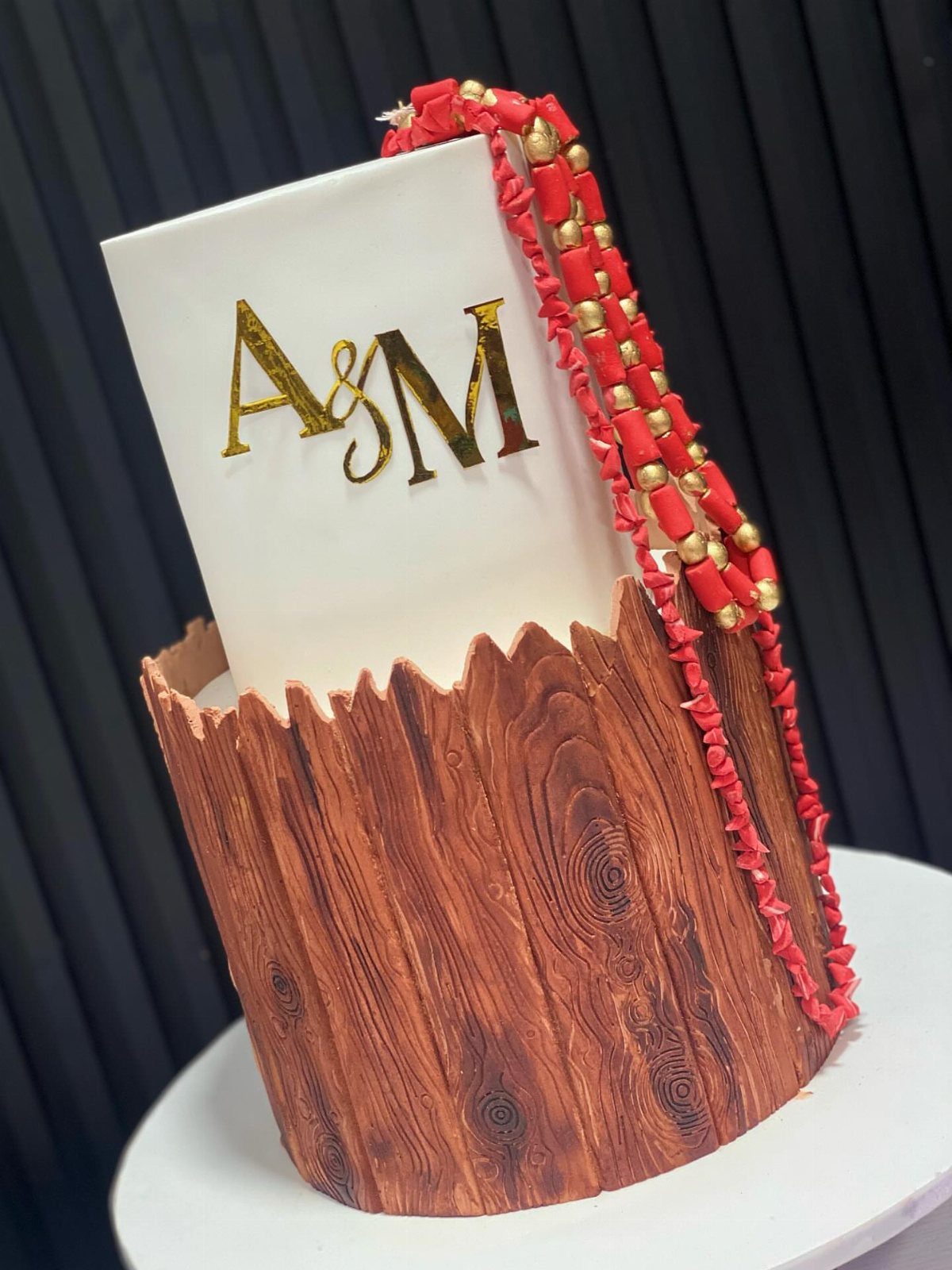 White cake with gold initials, wood-textured base, and red beaded necklaces decoration.