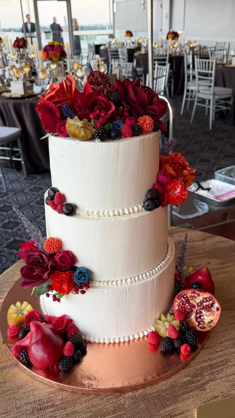 Three-tier white wedding cake decorated with red roses, blackberries, and pomegranate seeds.