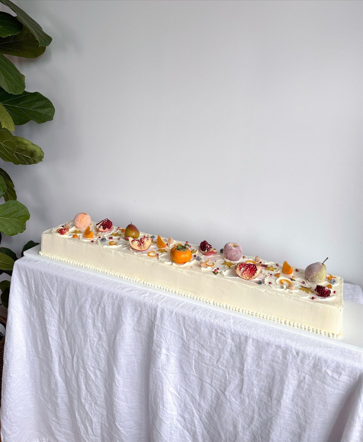 Long white frosted cake topped with fresh autumn fruits on a white tablecloth.