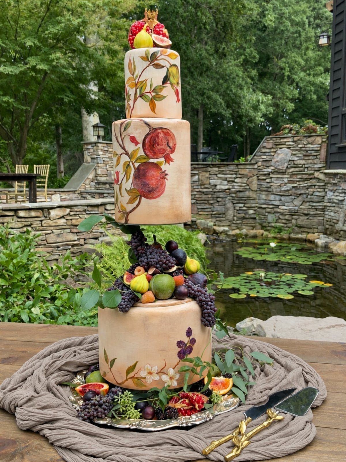 Three-tier wedding cake decorated with painted fruit and real grapes and pomegranates outdoors.