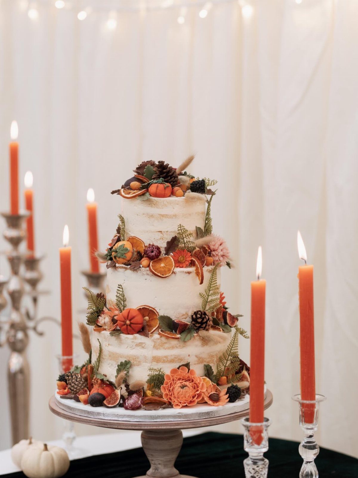 Three-tier semi-naked cake decorated with pumpkins, autumn fruits, and orange flowers.