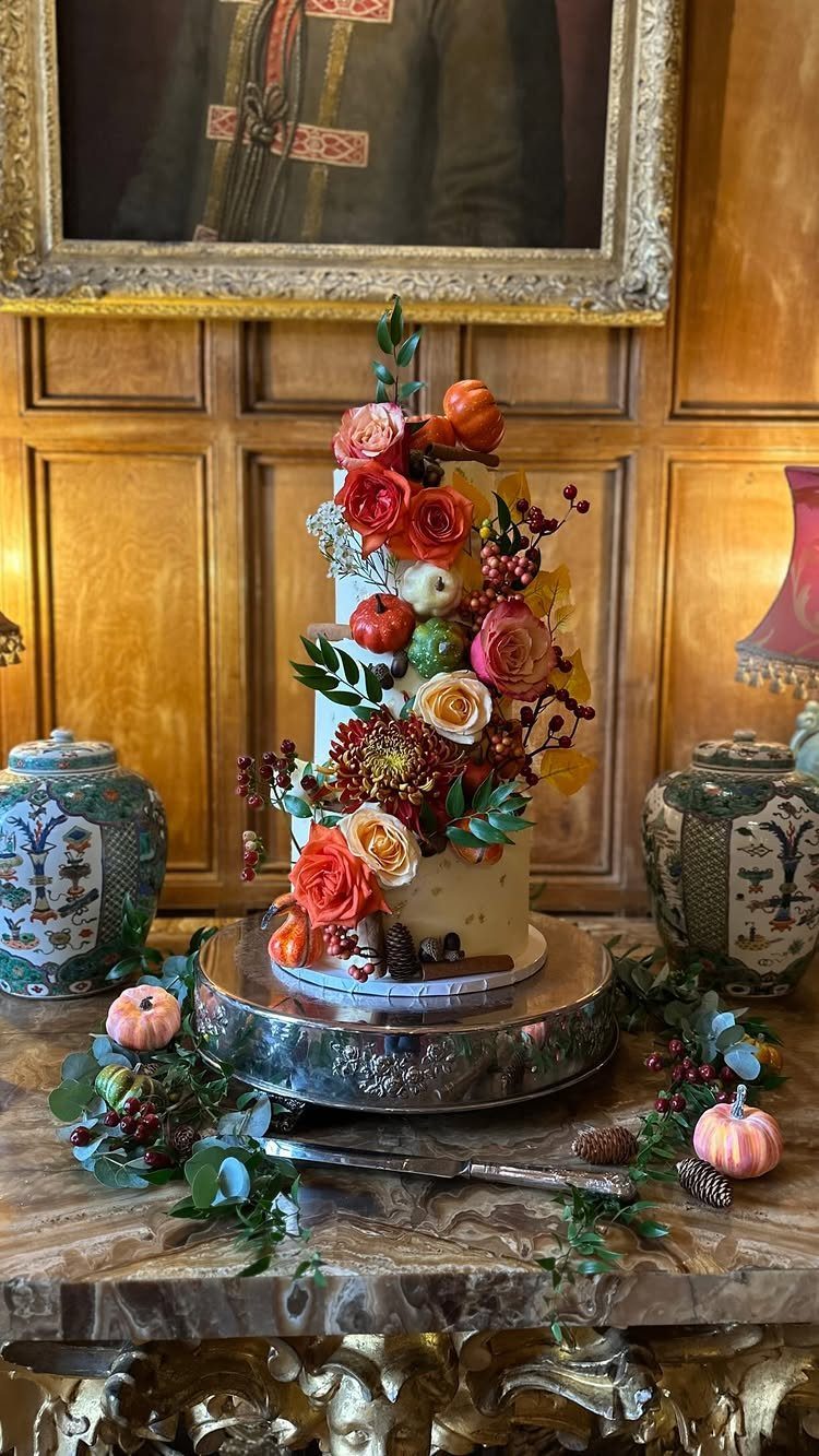 Multi-tiered white cake decorated with autumn flowers and pumpkins on a silver stand.