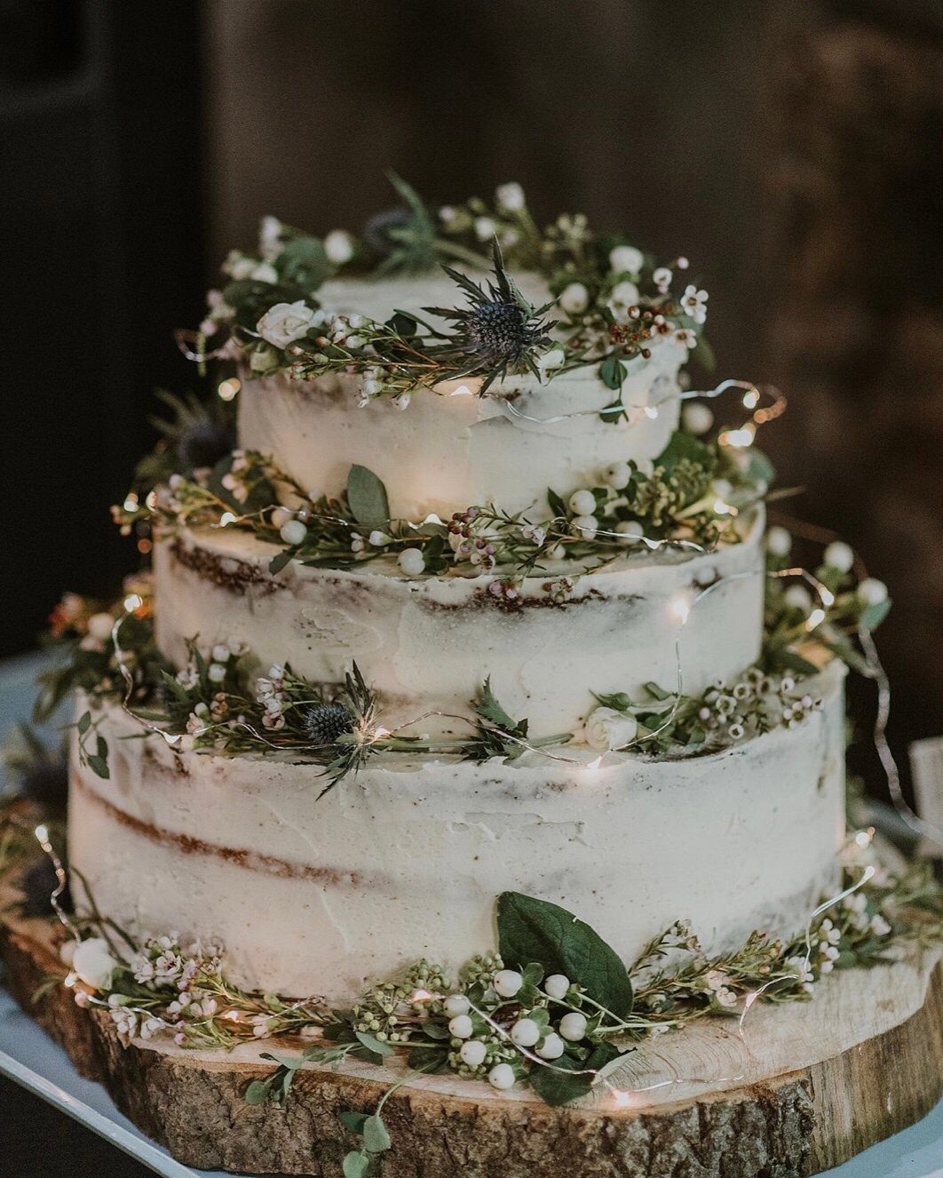 Three-tier semi-naked wedding cake decorated with greenery and thistles on a wood slice.
