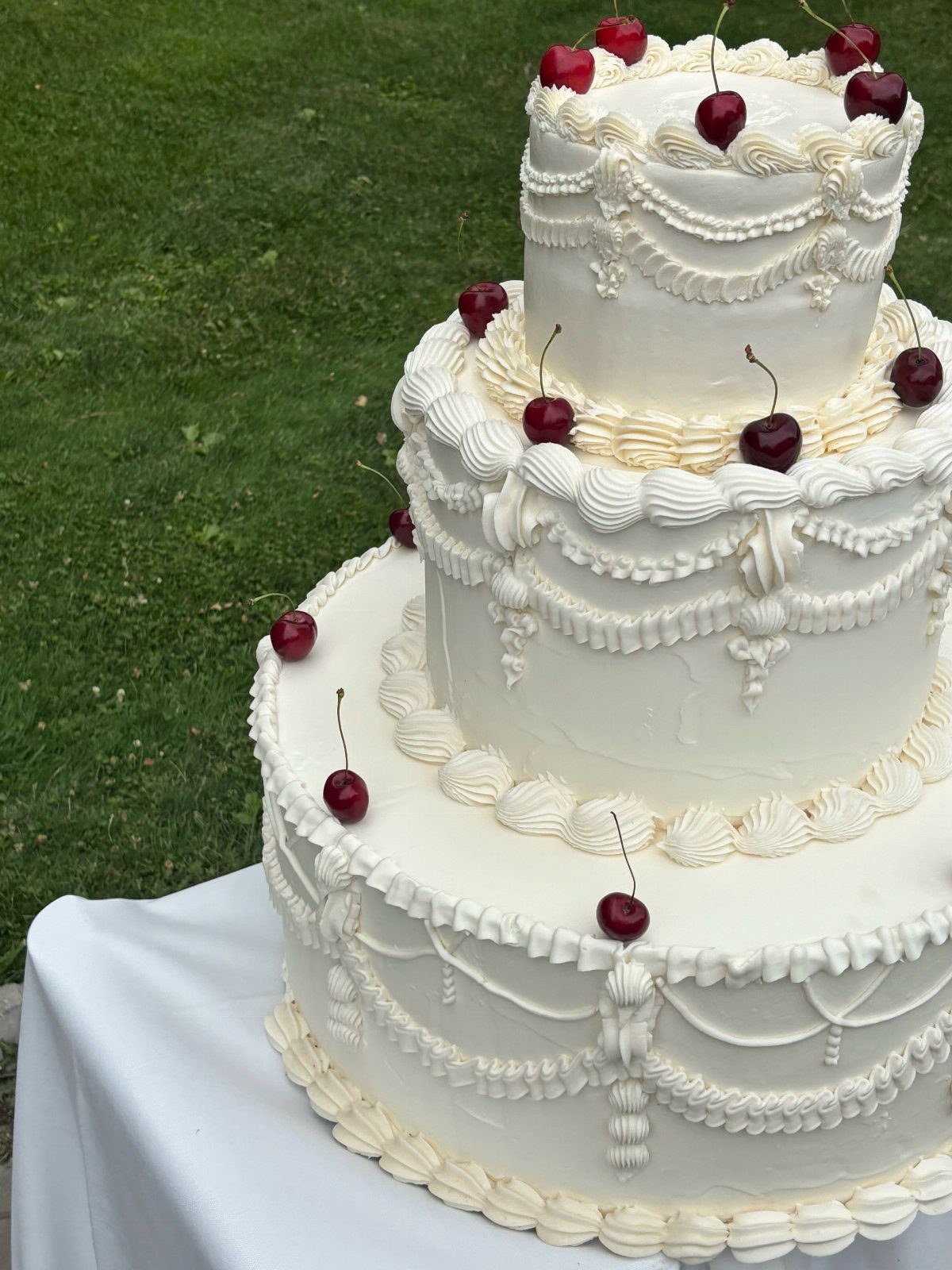Three-tier white wedding cake with buttercream piping and fresh red cherries on grass.